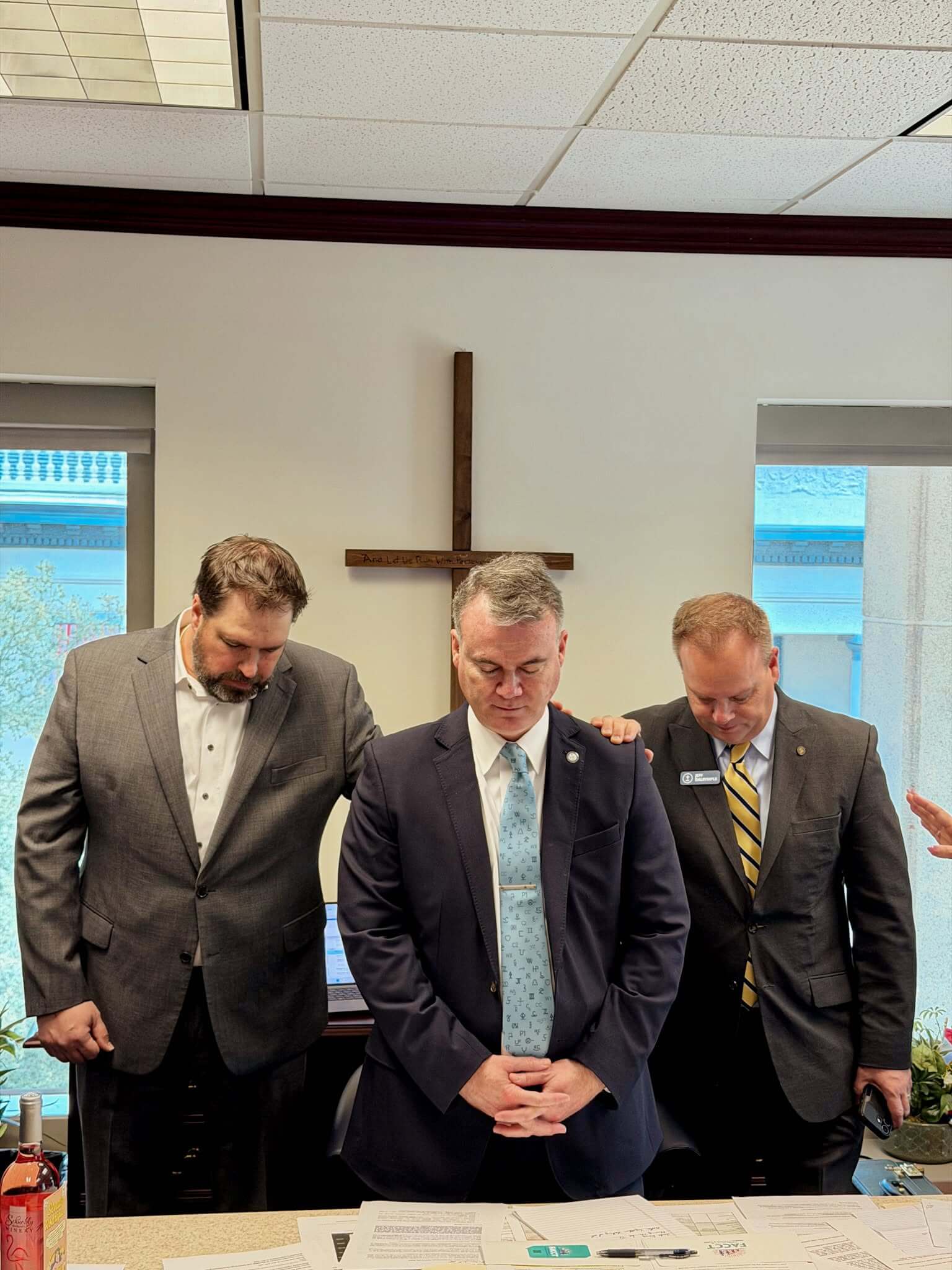 Three men in suits stand close together in an office, two with hands on the center man’s shoulders. The center man stands with eyes closed and head bowed. A large cross hangs on the wall behind them.