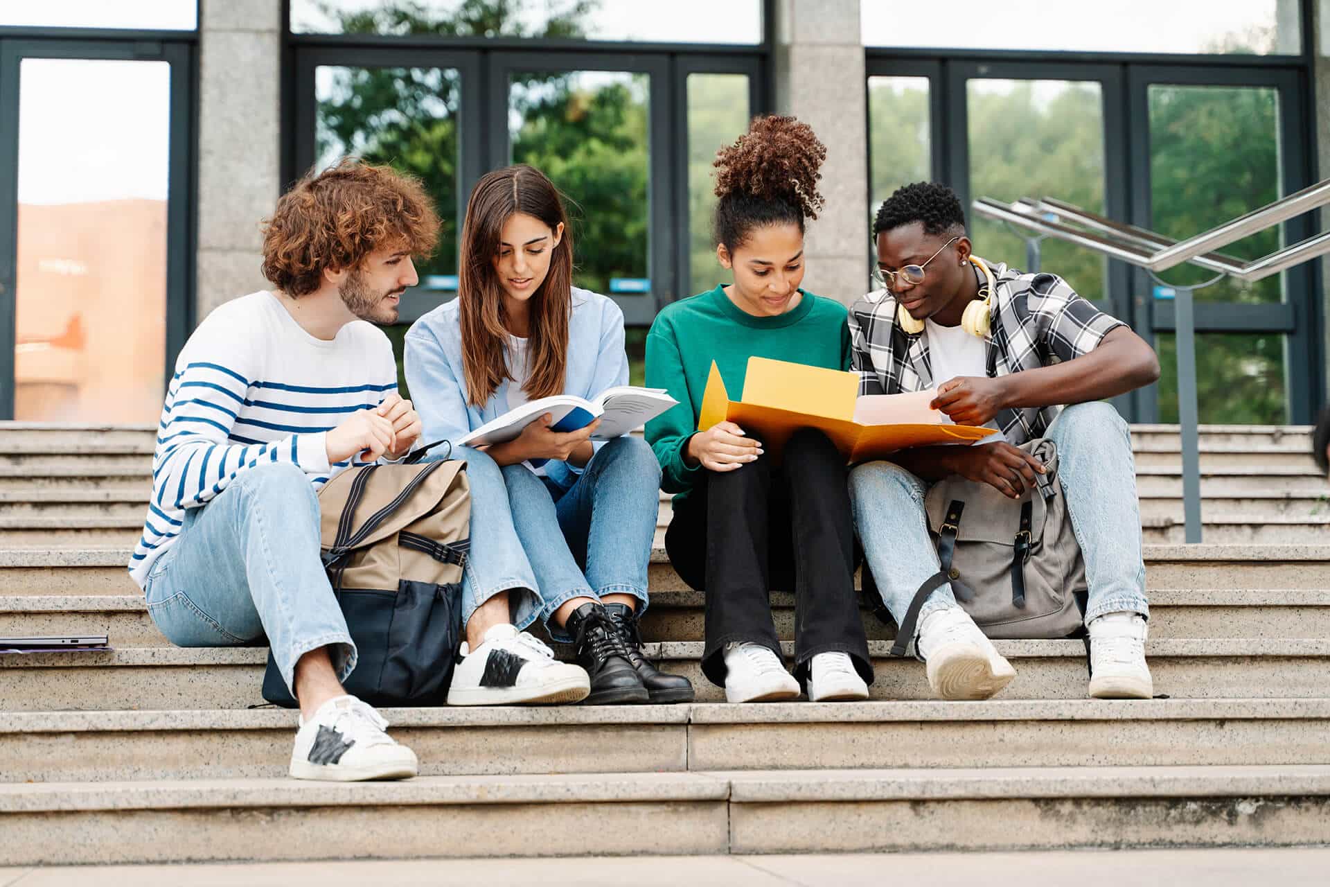 Four students sit on outdoor steps, reading books and notes together. They appear focused and collaborative, with backpacks beside them and a building entrance in the background.