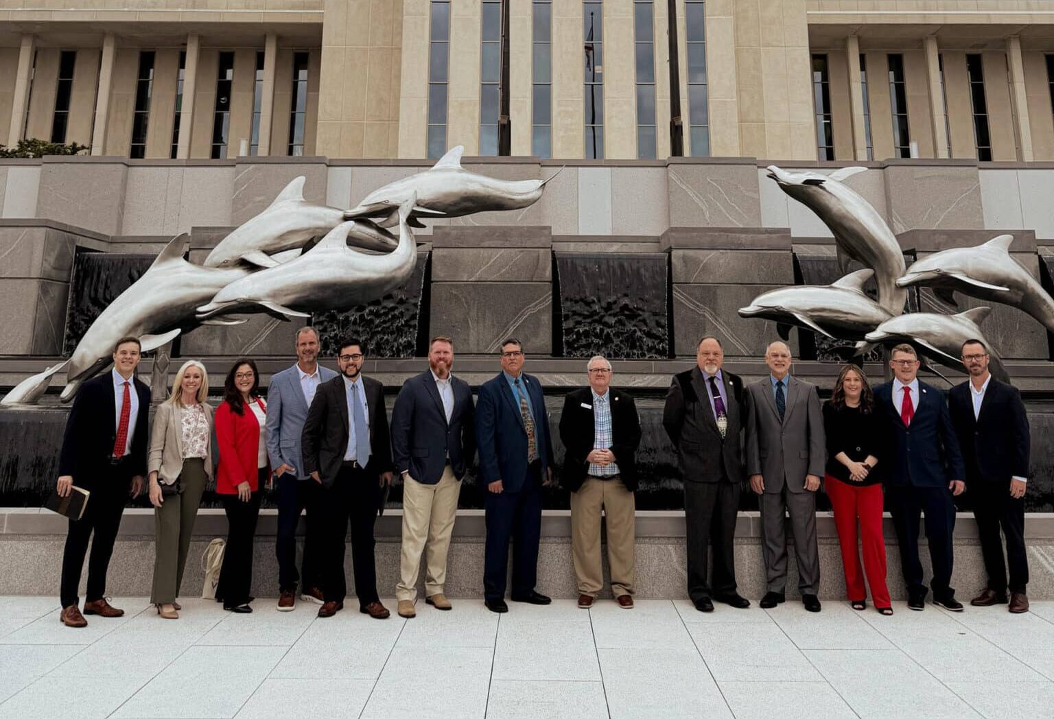 Thirteen people in business attire stand in front of Florida State Capitol and a sculpture of dolphins leaping over cascading water.