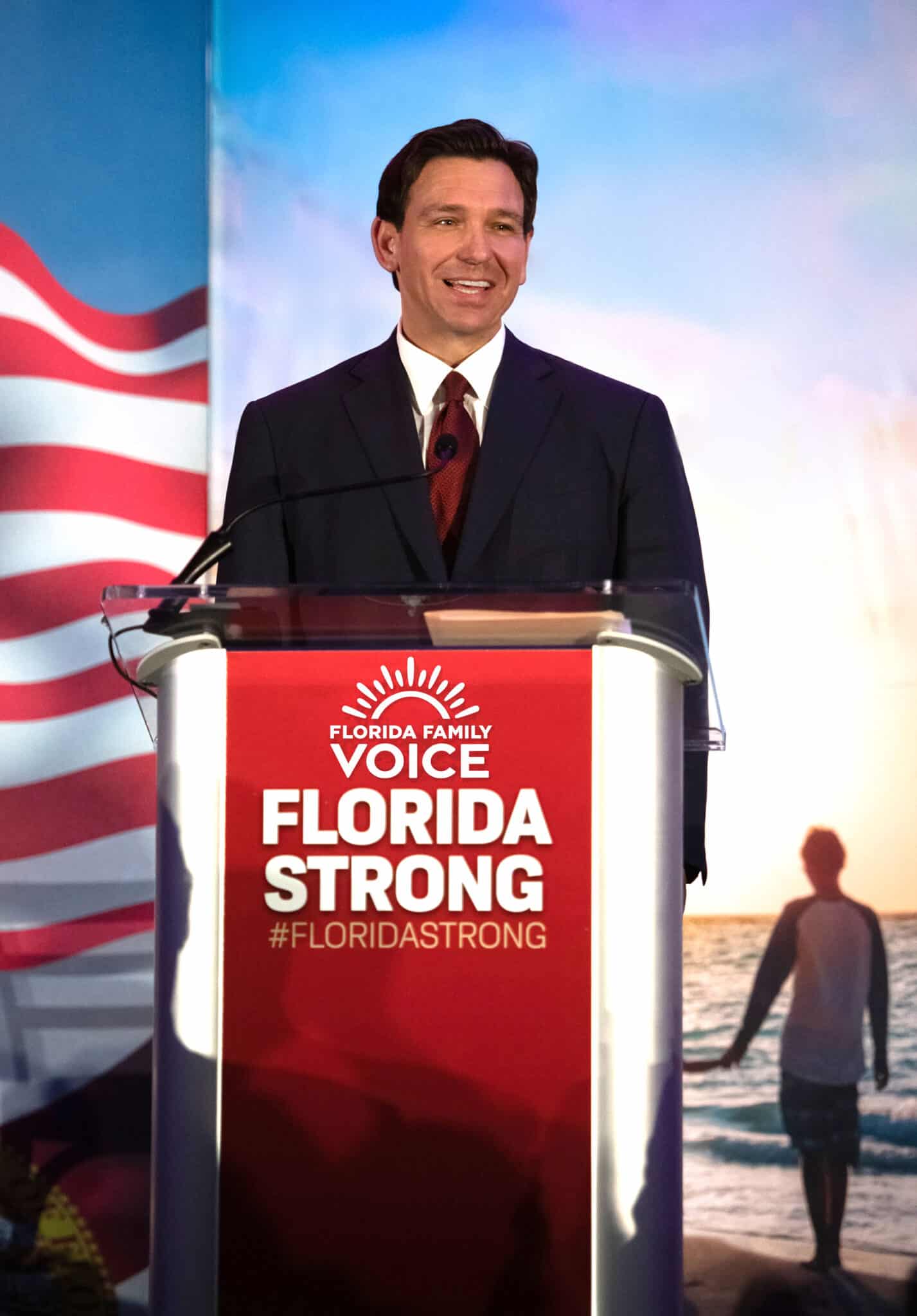 Governor Ron DeSantis speaks at a podium labeled “FLORIDA FAMILY VOICE FLORIDA STRONG #FLORIDASTRONG,” with an American flag and beach scene in the background.