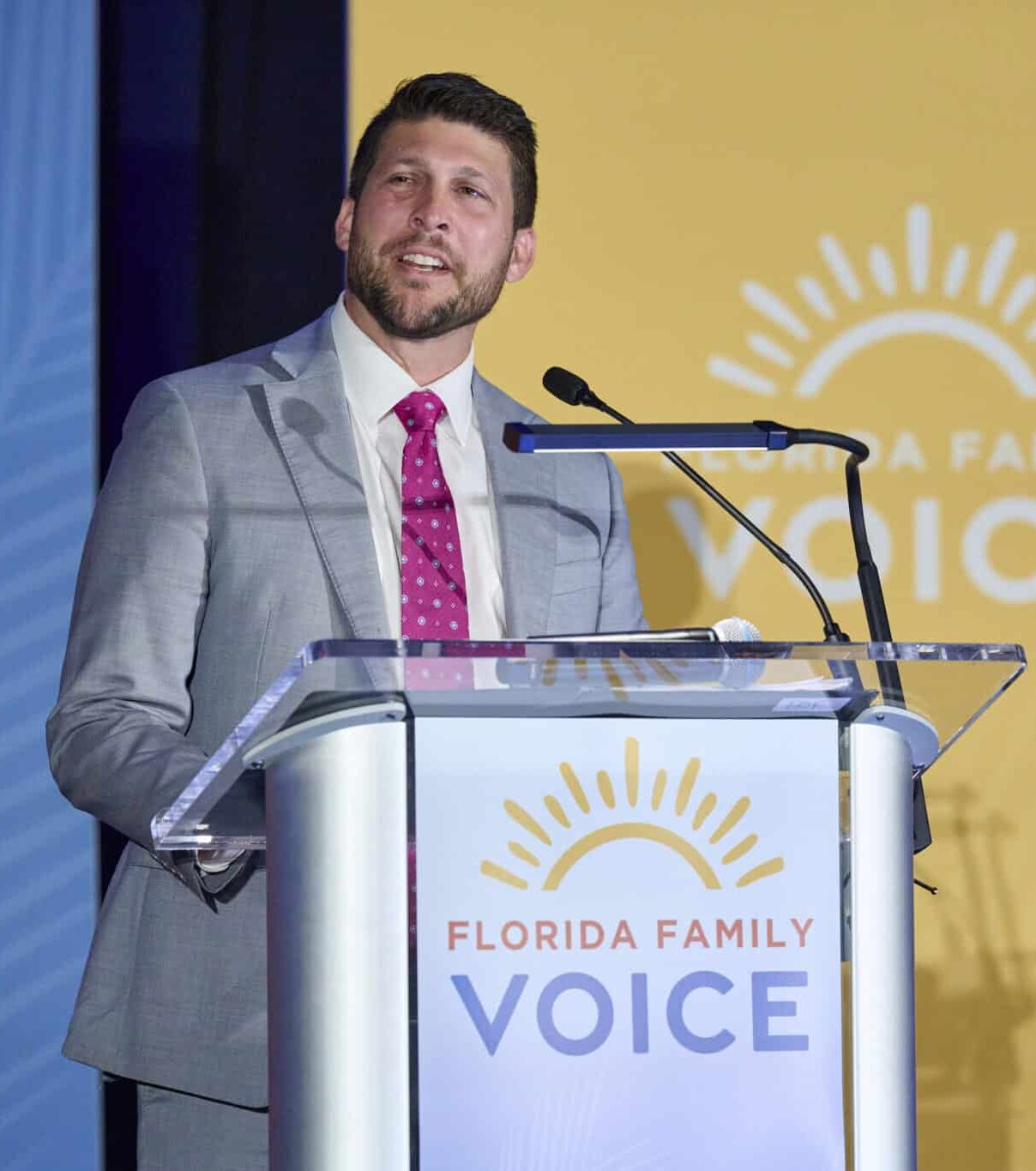 James Uthmeier in a gray suit and pink tie speaks at a podium labeled “Florida Family Voice,” with a bright yellow background featuring a sun graphic and the same text.
